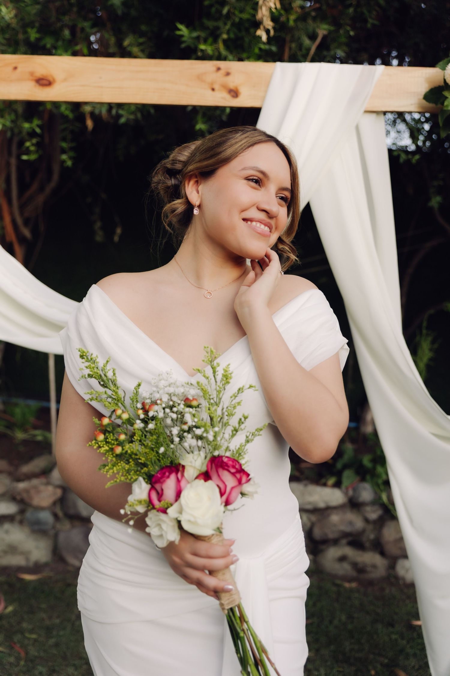 Smiling bride in white off-shoulder gown holding colorful bouquet standing before wooden arch with white draping outdoors.
