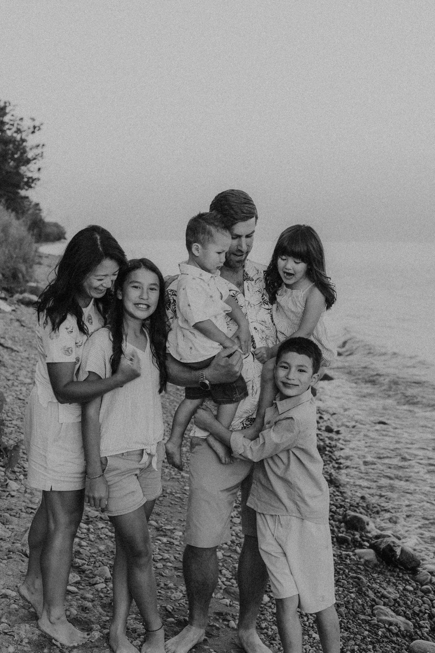 Black and white family photo of parents with four children smiling together on a rocky beach shoreline.