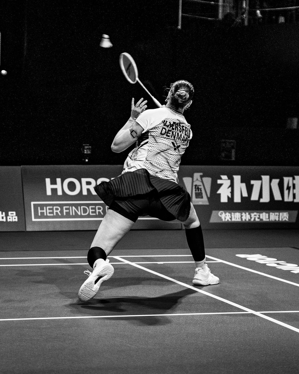 Black and white photo of a badminton player mid-swing on an indoor court during a competitive match.