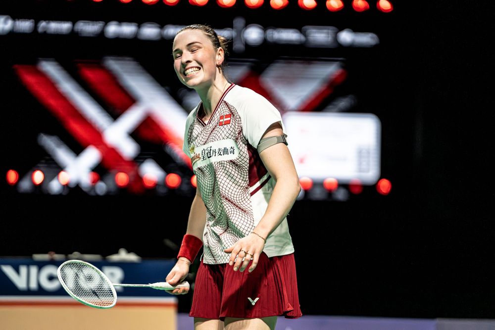 Female badminton player in red uniform smiling on court, holding racket during a professional tournament match.