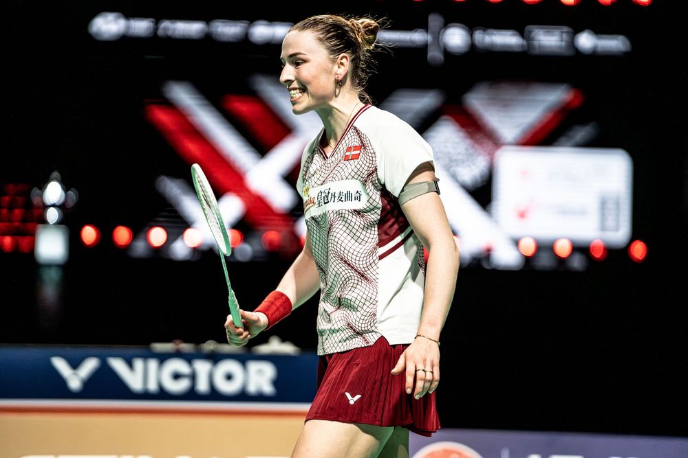 Female badminton player smiling on court at VICTOR tournament, holding racket in white and red sports uniform.