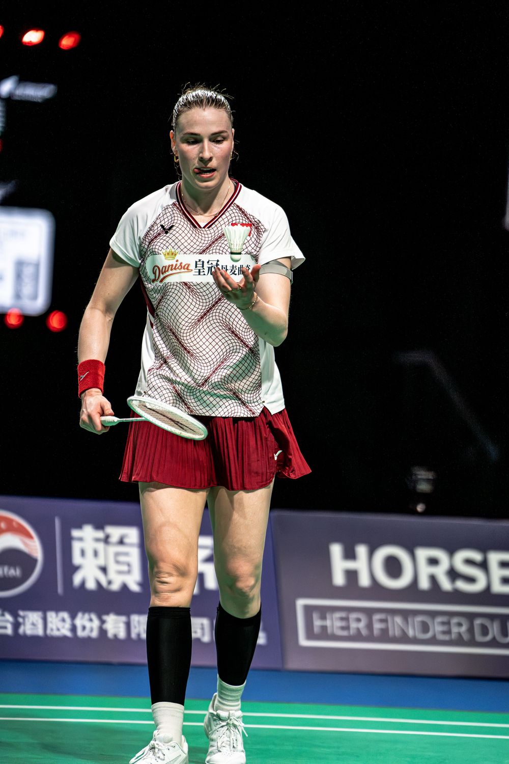 Female badminton player in white and red uniform holds racket on indoor court during competitive match.