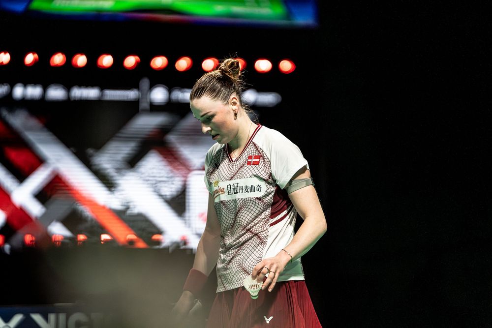 Female badminton player in red Victor jersey bowing her head during a competitive match under dramatic stage lighting.