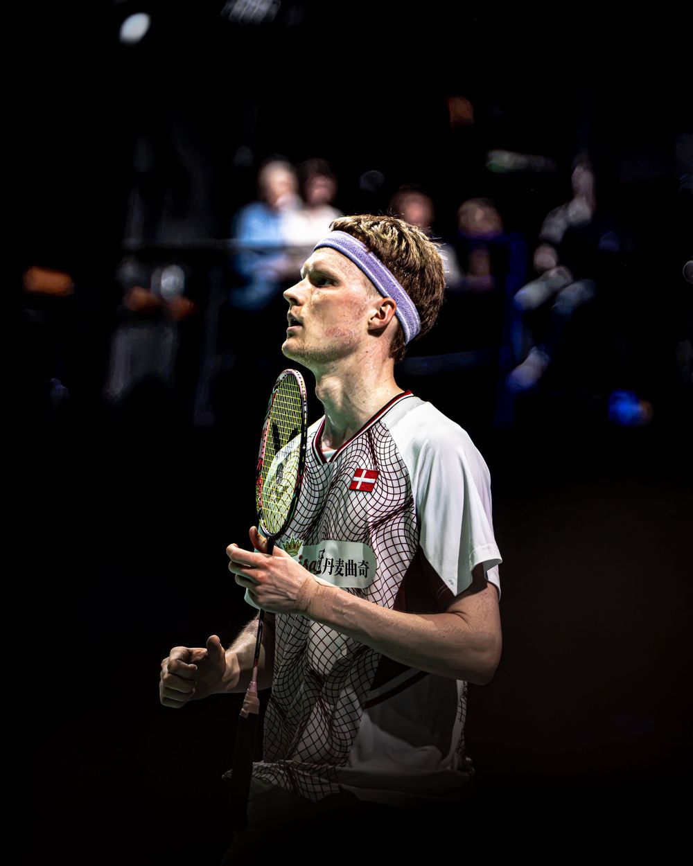 Male badminton player holding racket wearing purple headband and Denmark jersey competing in indoor arena.