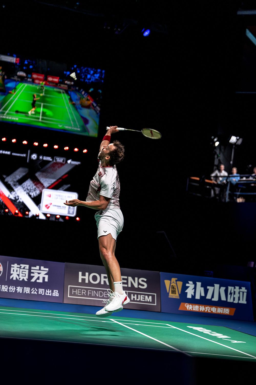 Professional badminton player jumping to smash shuttlecock during an indoor tournament match with large screen backdrop.