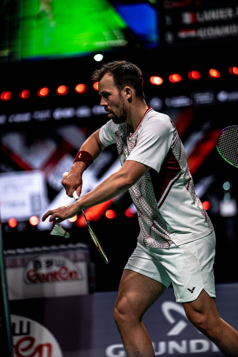 A male badminton player in white athletic wear executes a powerful shot with his racket during a professional tournament.