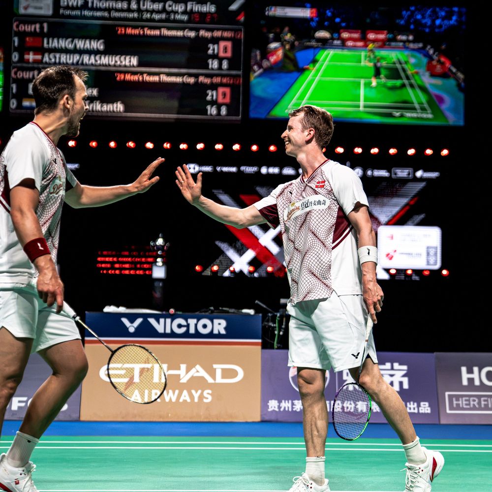 Two male badminton players high-fiving during BWF Thomas Cup Finals at VICTOR-sponsored indoor arena.