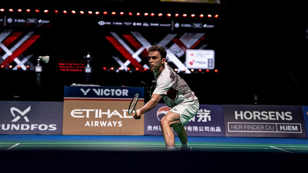 Male badminton player prepares to return a shot during a professional tournament at an indoor court.