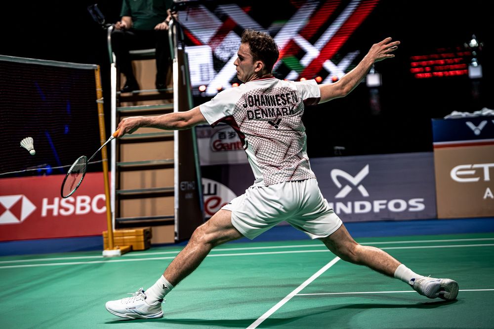 Male badminton player from Denmark lunges wide to return a shot during a professional indoor tournament.
