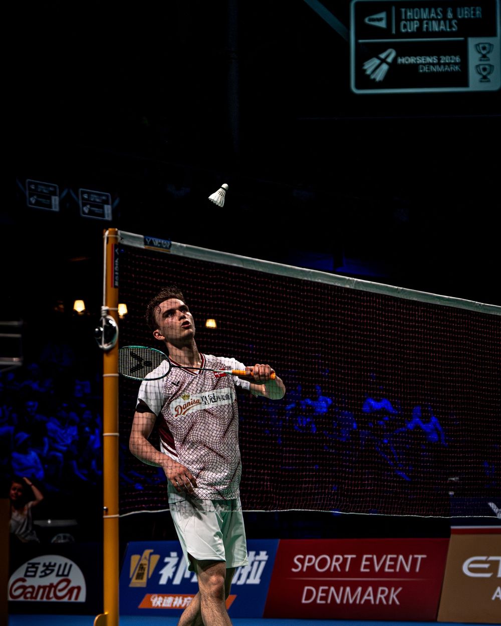Badminton player mid-swing at Thomas & Uber Cup Finals 2020 in Denmark, reaching for shuttlecock above the net.