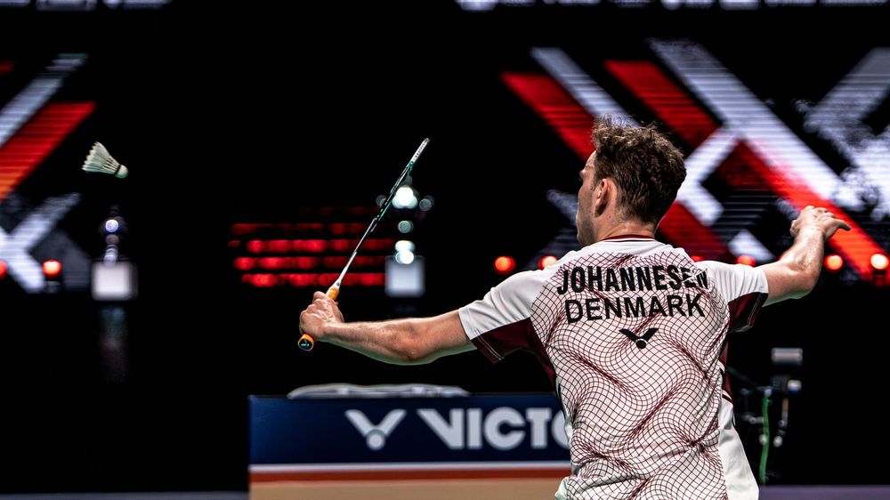 Danish badminton player Johannesen mid-swing at a Victor-sponsored tournament, viewed from behind in a packed arena.