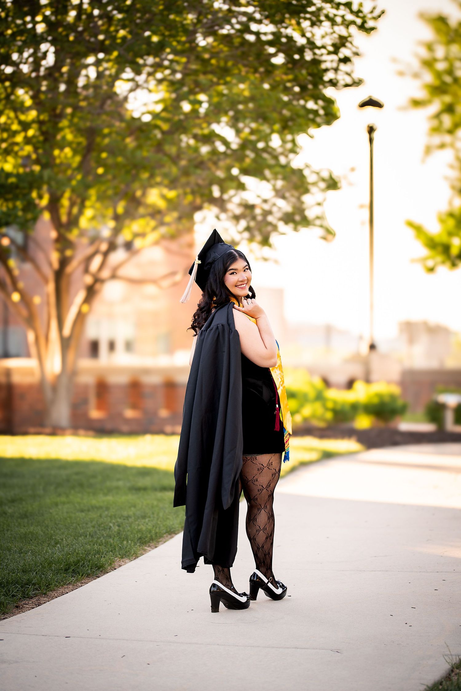 Graduate in black cap and gown walking on sunny campus sidewalk, glancing back.