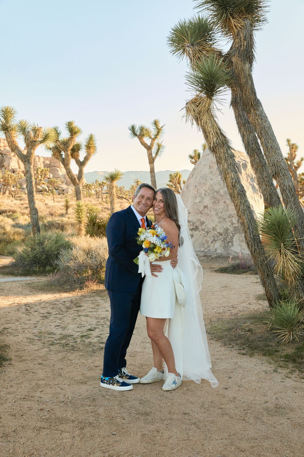 Bride and groom embrace in Joshua Tree desert landscape, she holds colorful bouquet, both wearing sneakers.