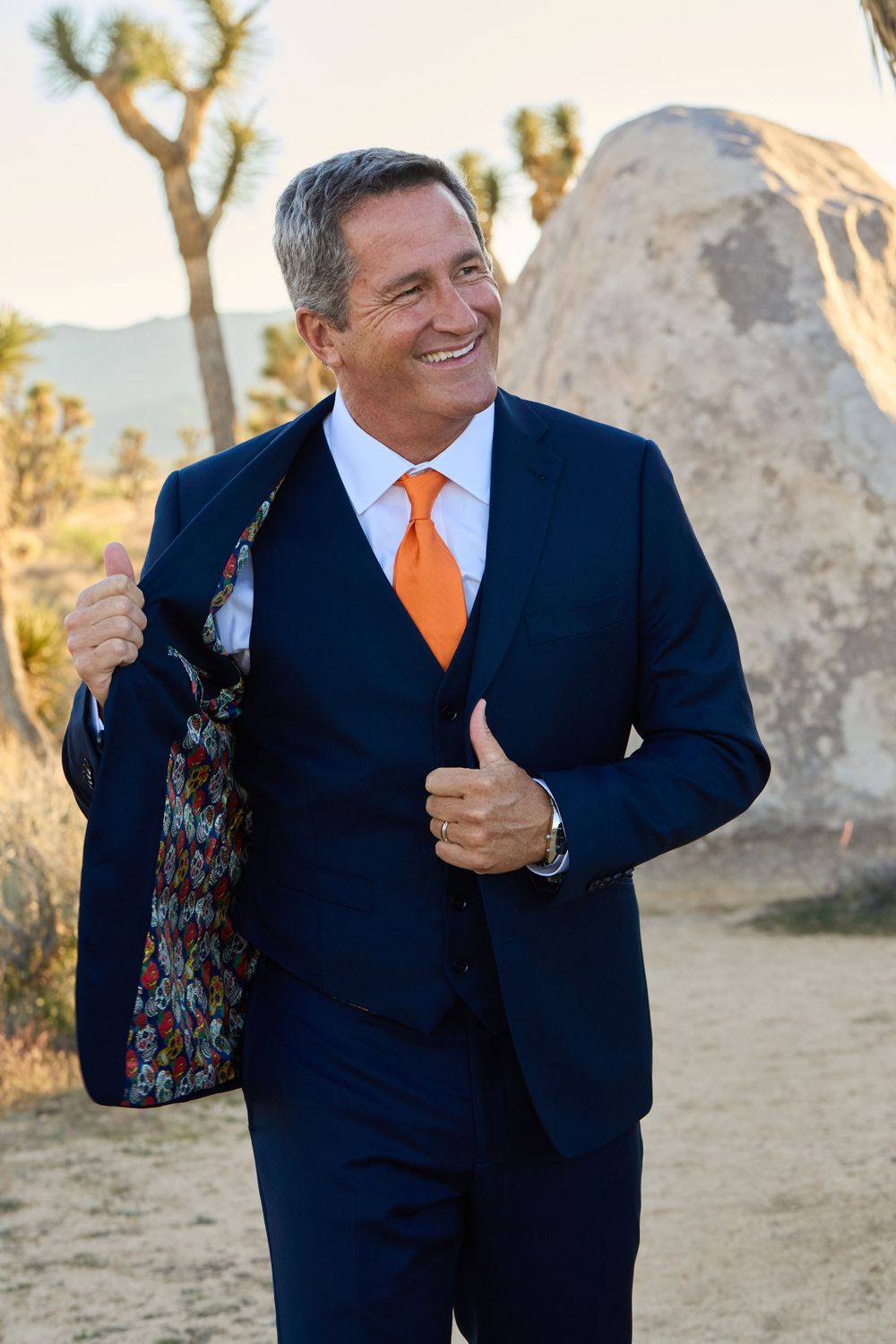 Smiling man in navy blue suit with orange tie and floral lining poses outdoors in desert landscape.
