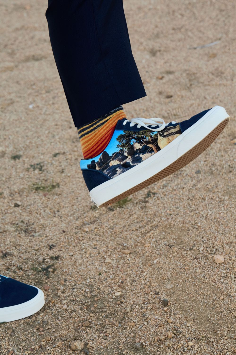 Person wearing navy blue high-top sneakers with tropical print and colorful striped socks, stepping on sandy ground.