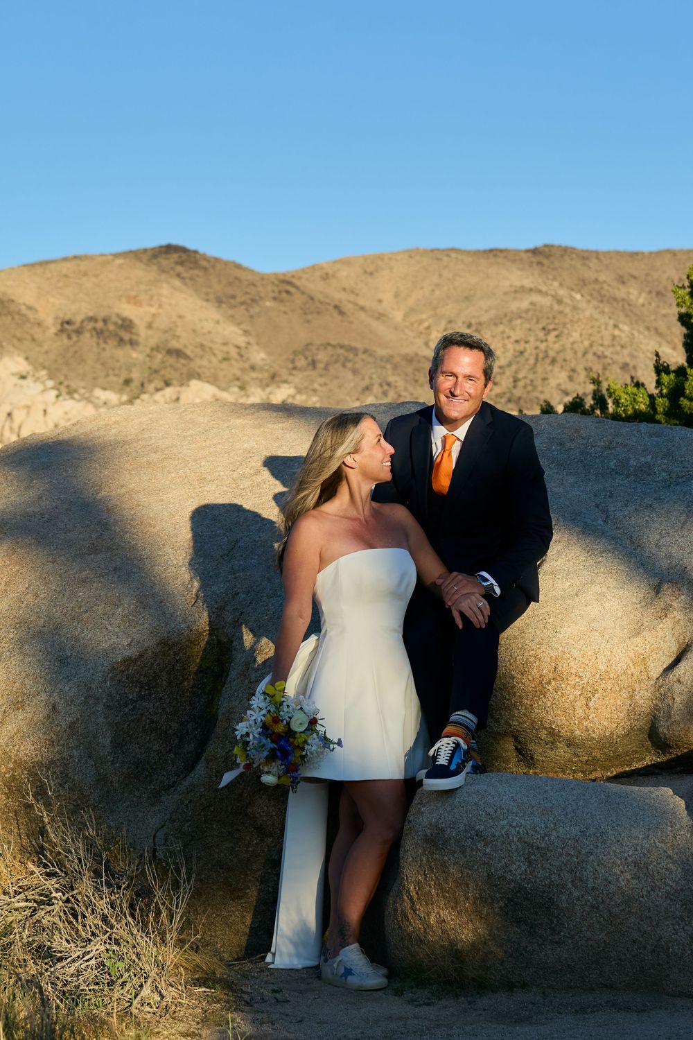 Bride in white strapless dress and groom in navy suit with orange tie pose on desert rocks with mountains behind.