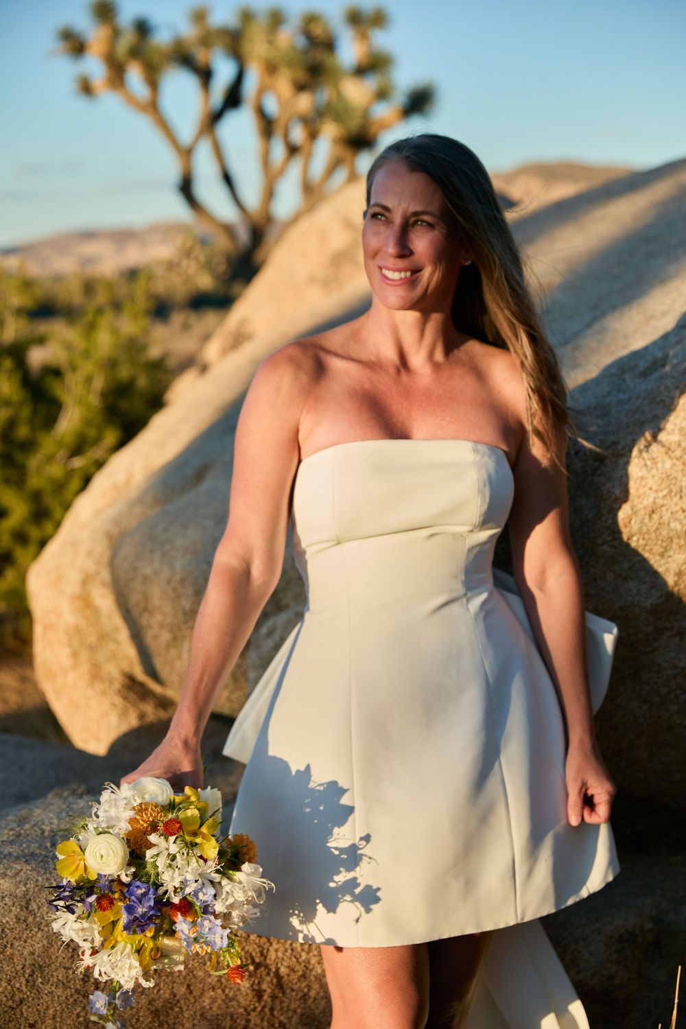Smiling bride in white strapless mini dress holding wildflower bouquet among Joshua Tree rocks at golden hour.