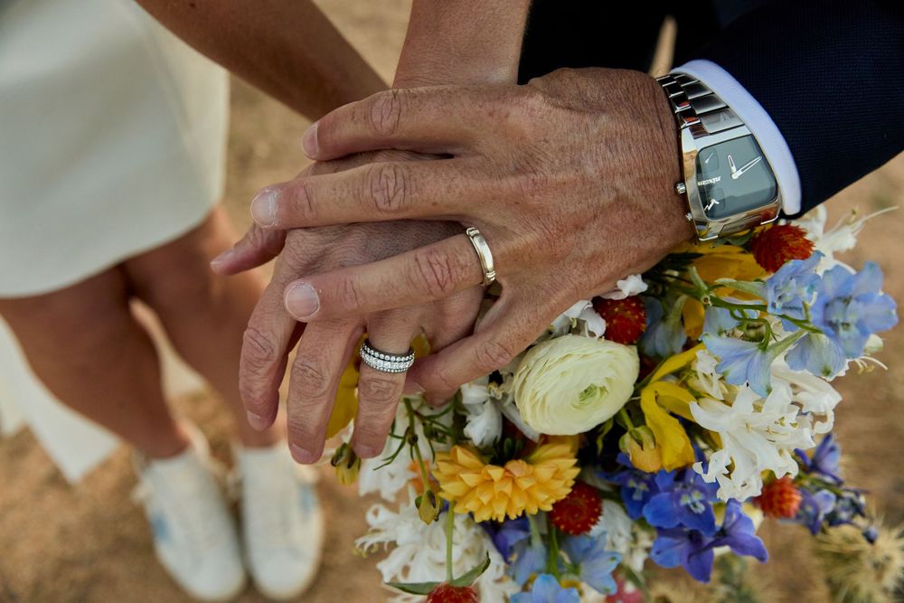 Newlyweds holding hands showing wedding rings resting on a colorful bridal bouquet of wildflowers.