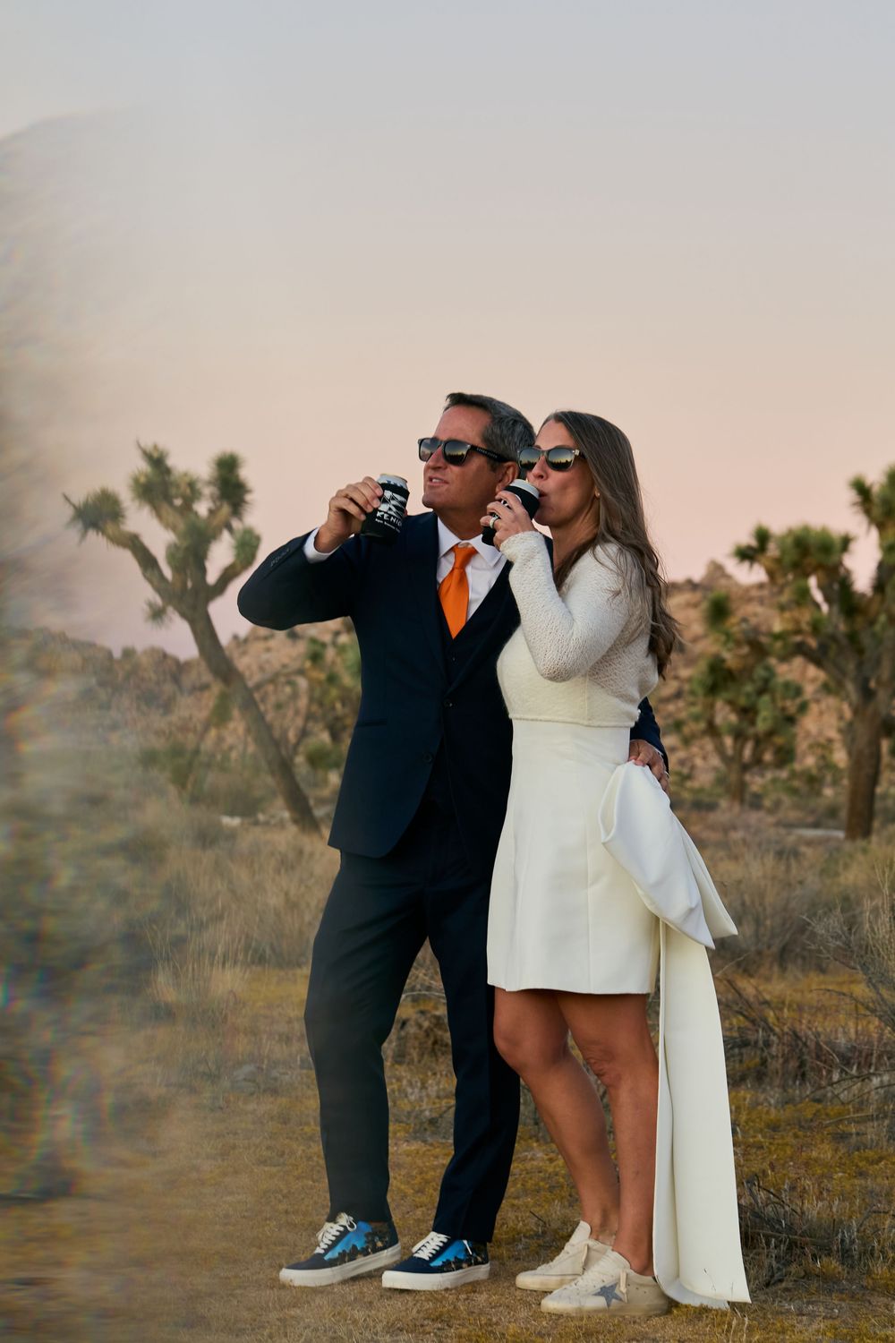 Couple in wedding attire drinking canned beverages among Joshua trees in desert landscape at sunset.