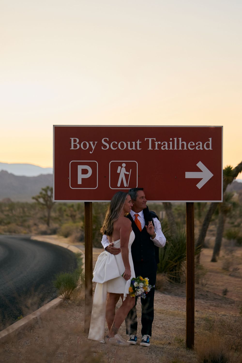 Bride and groom embrace under Boy Scout Trailhead sign at Joshua Tree National Park during golden hour sunset.