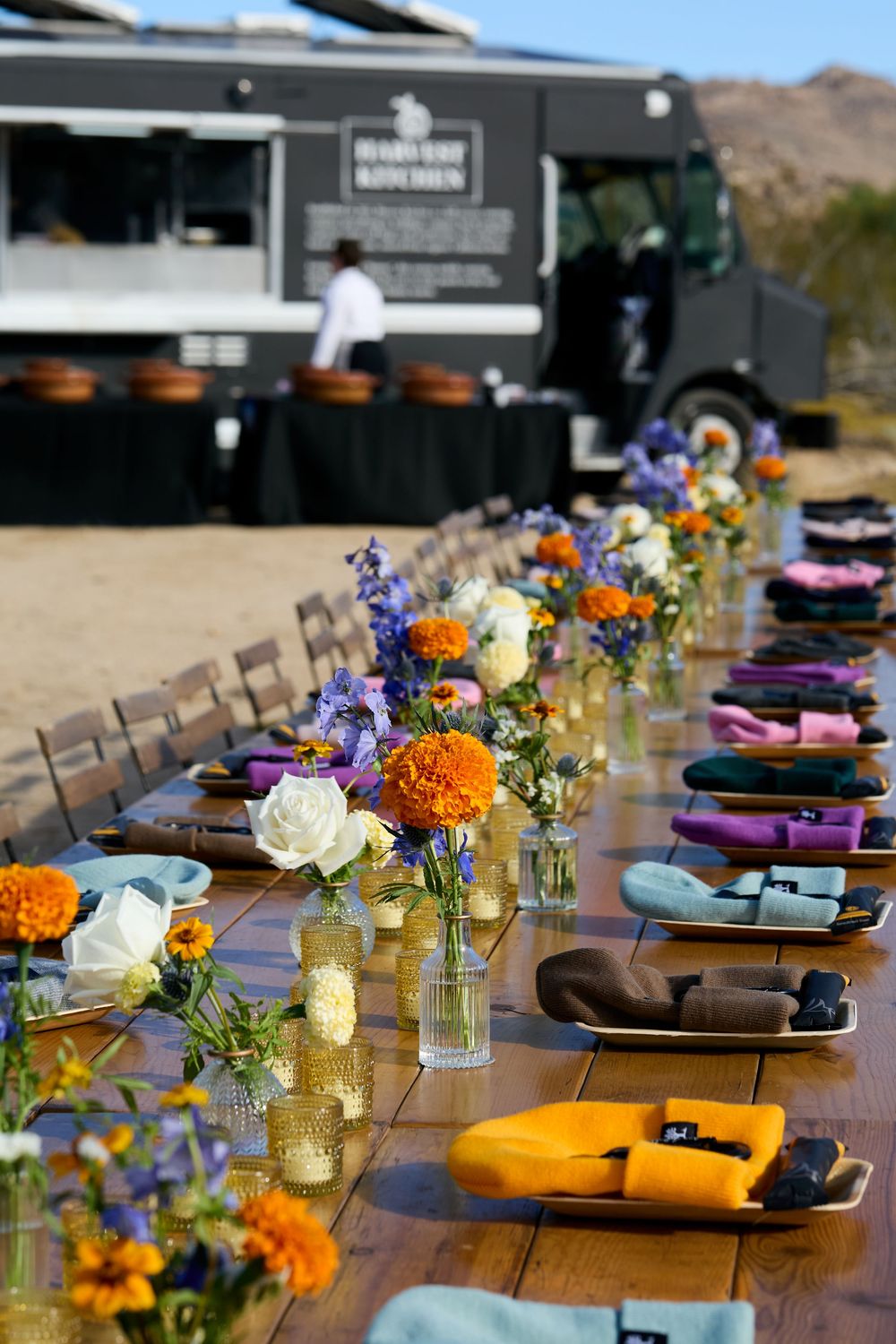 Rustic outdoor wedding reception table with colorful floral centerpieces and food truck in desert setting.