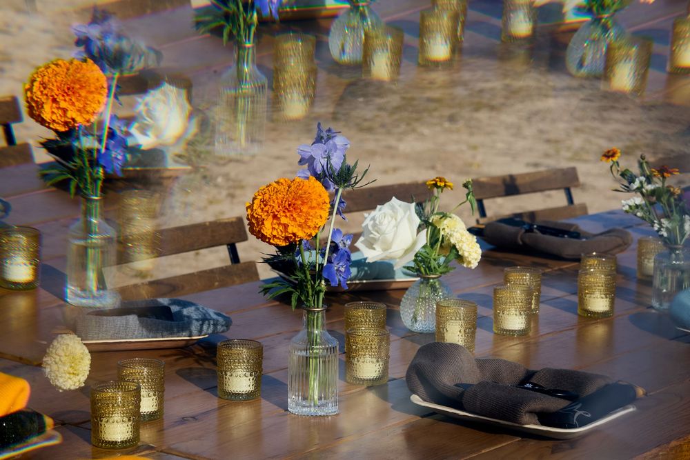 Rustic wedding reception table with orange marigolds, white roses, blue flowers, and gold votive candles as centerpieces.