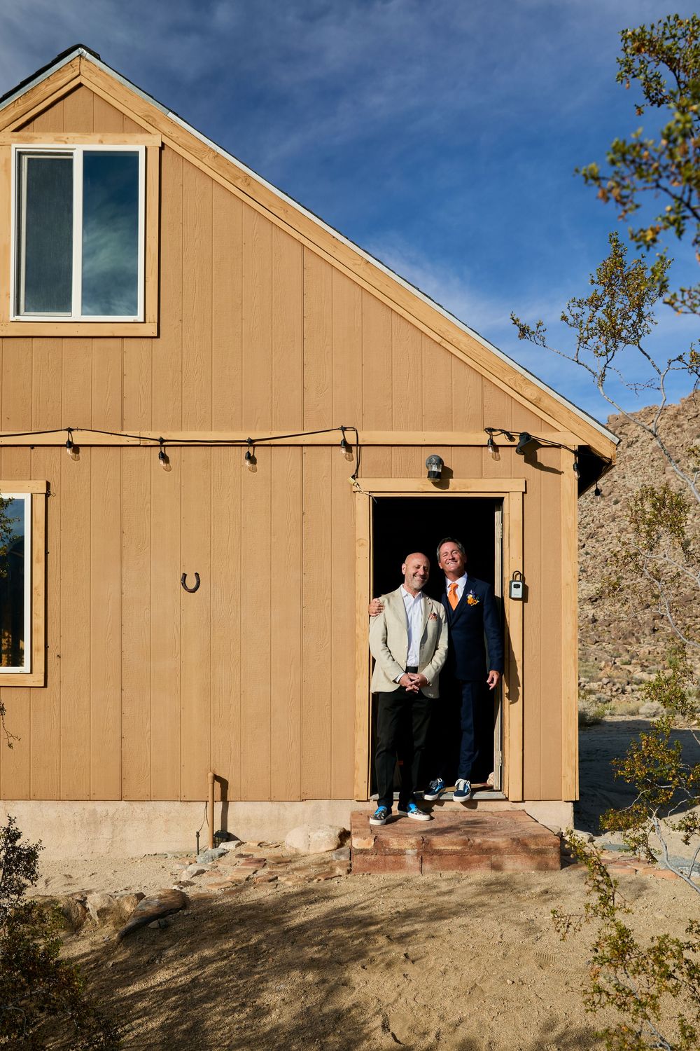 Two grooms pose together in the doorway of a rustic tan barn at their outdoor desert wedding.