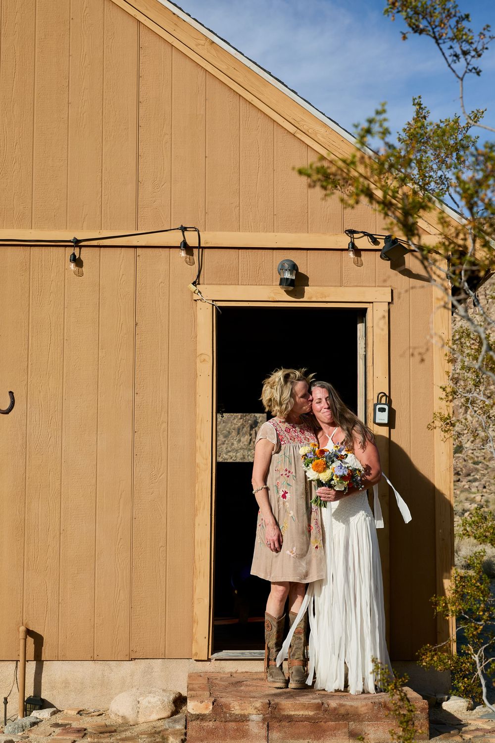 A bride in a white gown holds a bouquet while standing with a guest outside a rustic barn wedding venue.