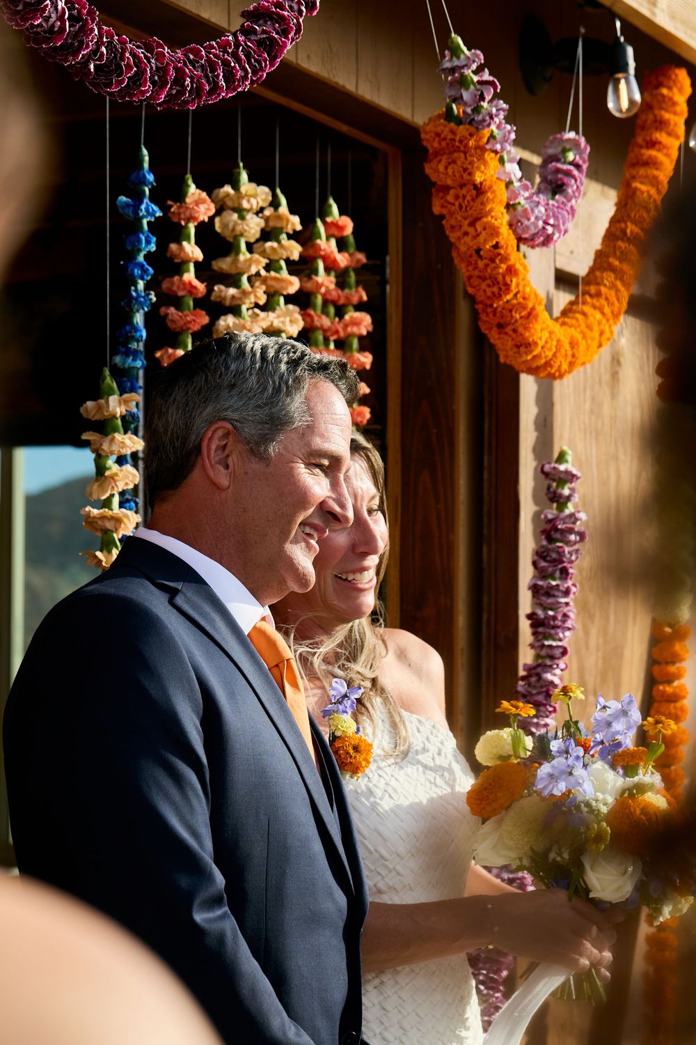 Smiling bride and groom stand beneath colorful floral garlands during an outdoor wedding ceremony.