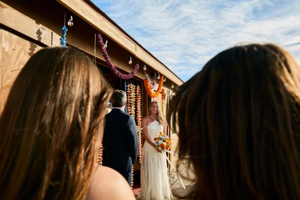 Wedding ceremony viewed from behind guests as bride and groom stand at colorful floral altar.