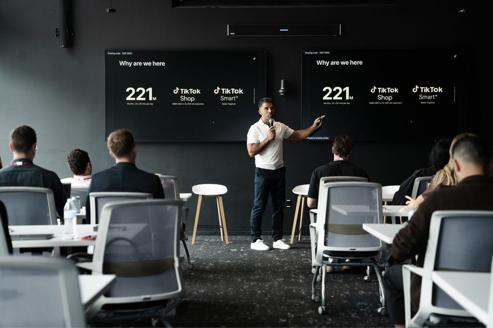 A presenter speaks at a TikTok Shop business conference, standing before a large screen displaying statistics in a modern dark meeting room.