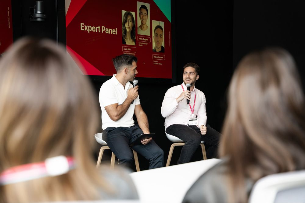 Two men speak on stage at an Expert Panel event, seated on chairs with a presentation screen behind them.