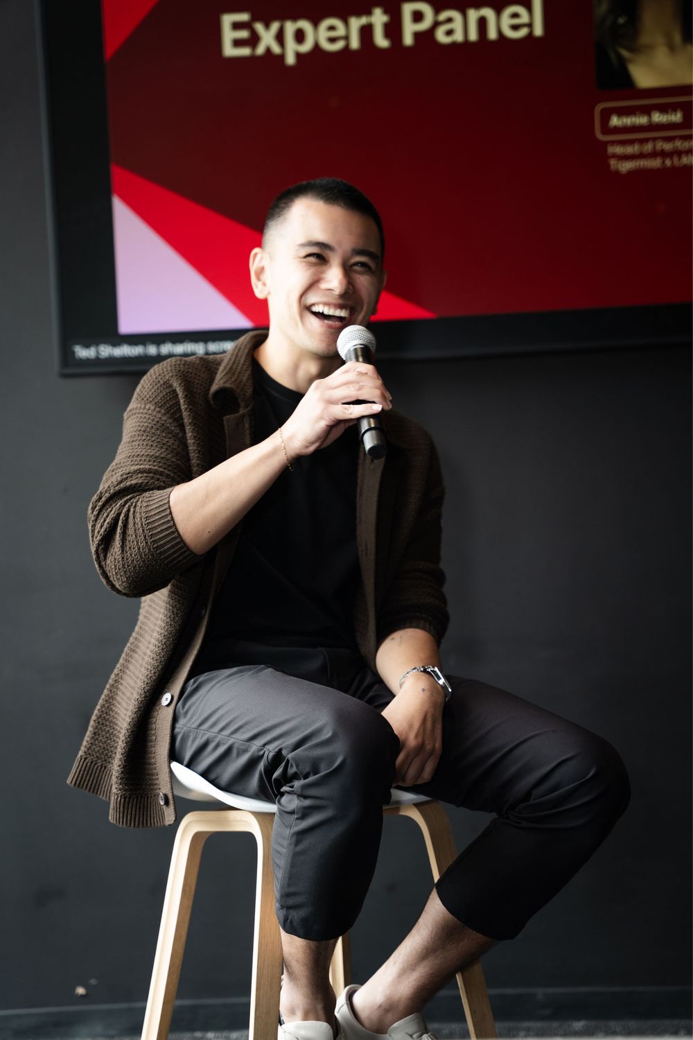 Young man smiling and holding microphone while seated on stool at Expert Panel event.