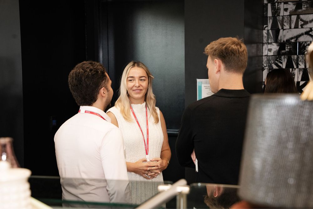 Three professionals networking at a conference, smiling and conversing near a reception desk.