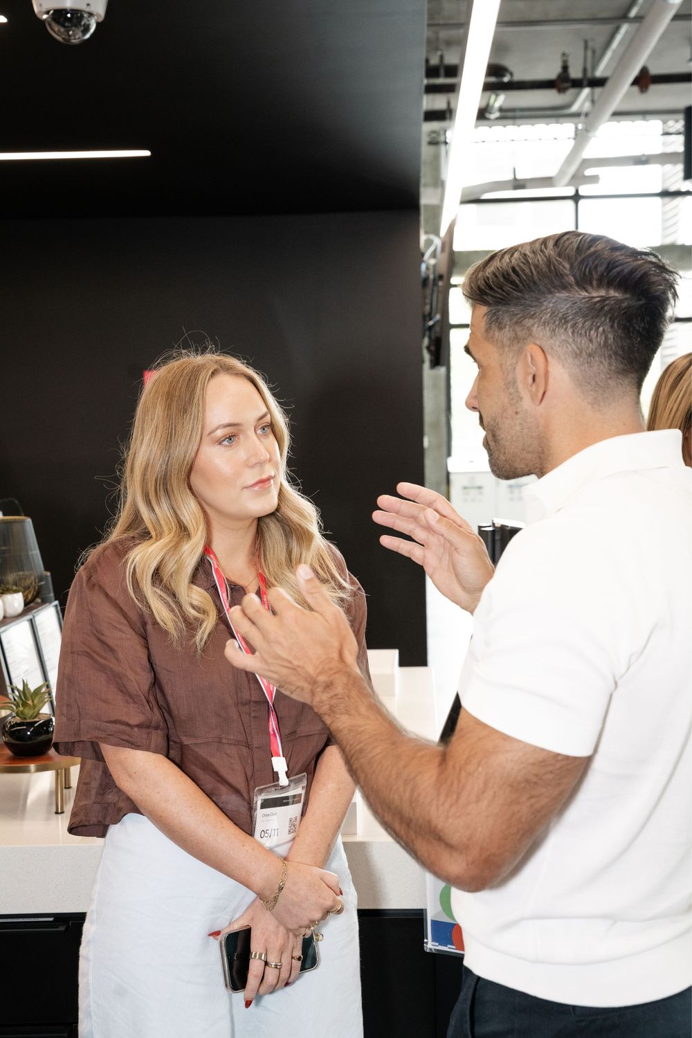 A blonde woman with a lanyard speaks with a man in a white polo shirt at a modern office reception desk.