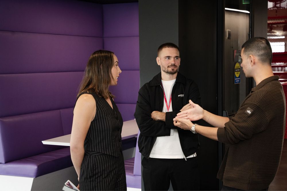 Three colleagues networking in a modern office lounge with purple booth seating during a business meeting.