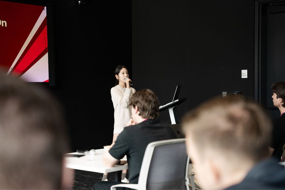 Woman presenting at a corporate conference in a modern dark auditorium with an engaged audience seated in the foreground.