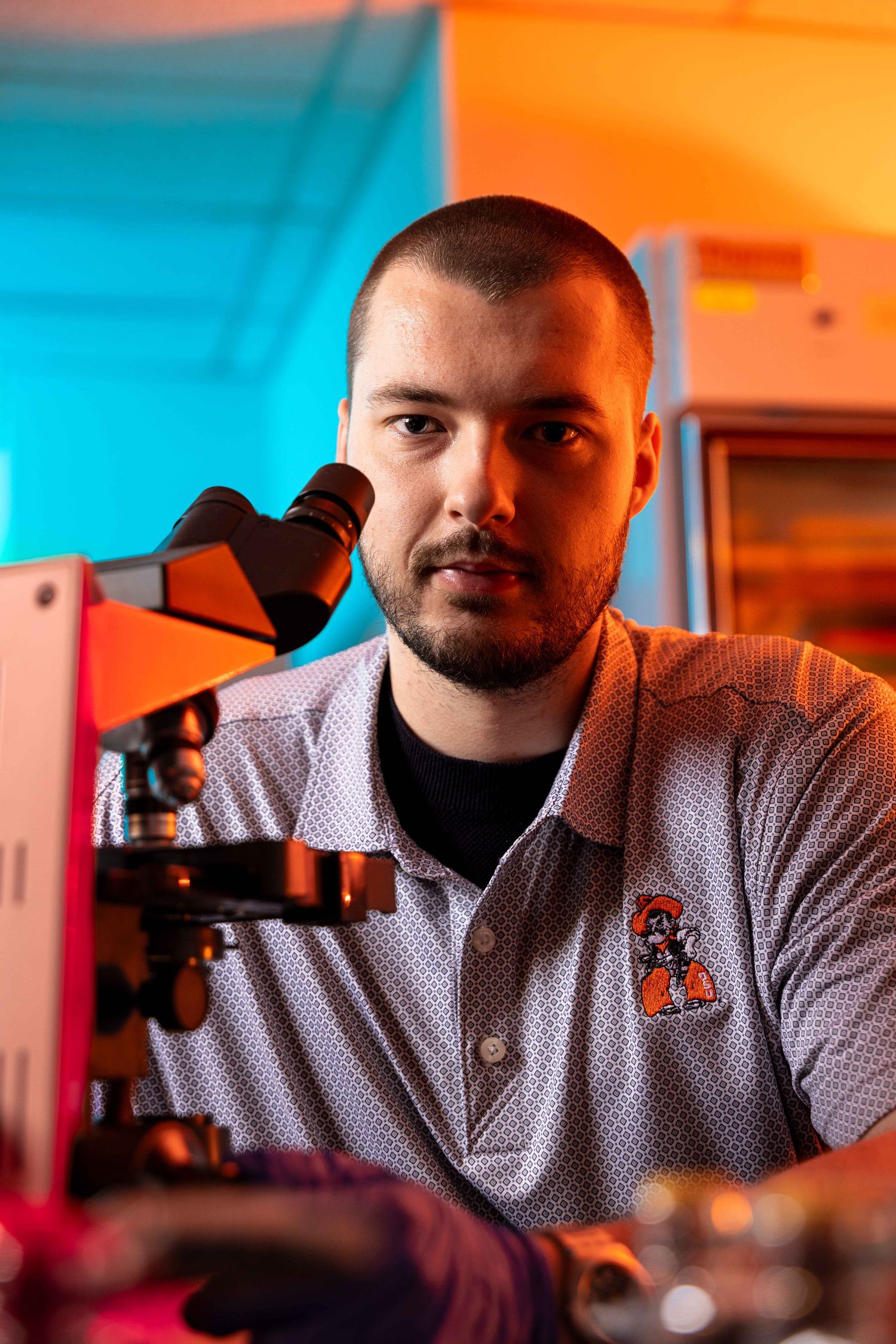 A young male scientist with a beard examines a microscope in a lab lit with colorful neon lighting.