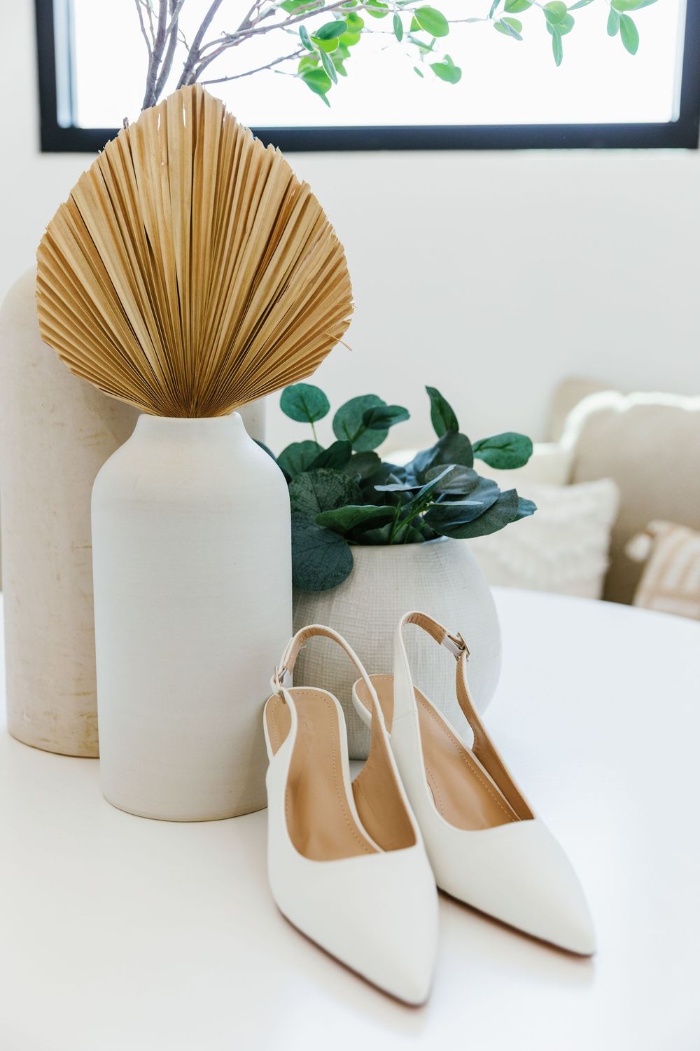 Elegant bridal shoe details featuring white pointed-toe slingback heels alongside neutral ceramic vases and dried palm decor.