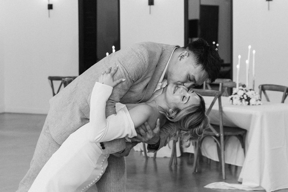 Black and white wedding photography of a groom dipping and kissing his bride during a first dance indoors.