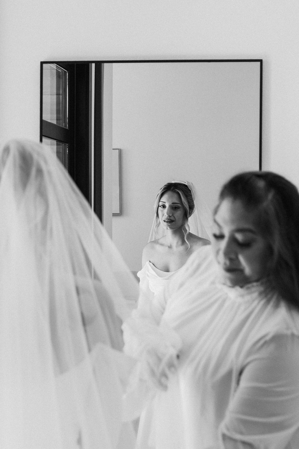 Black and white candid wedding preparation photo of a mother helping adjust her daughter's veil near a mirror.