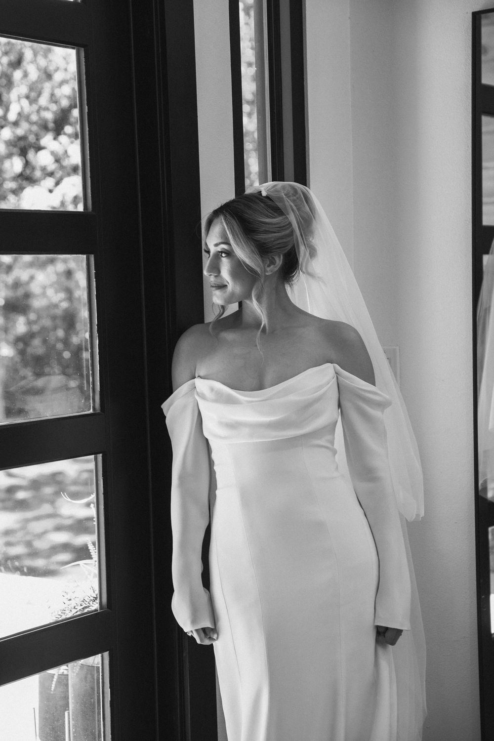 Black and white bridal portrait of a bride in an off-the-shoulder gown standing beside a window indoors.