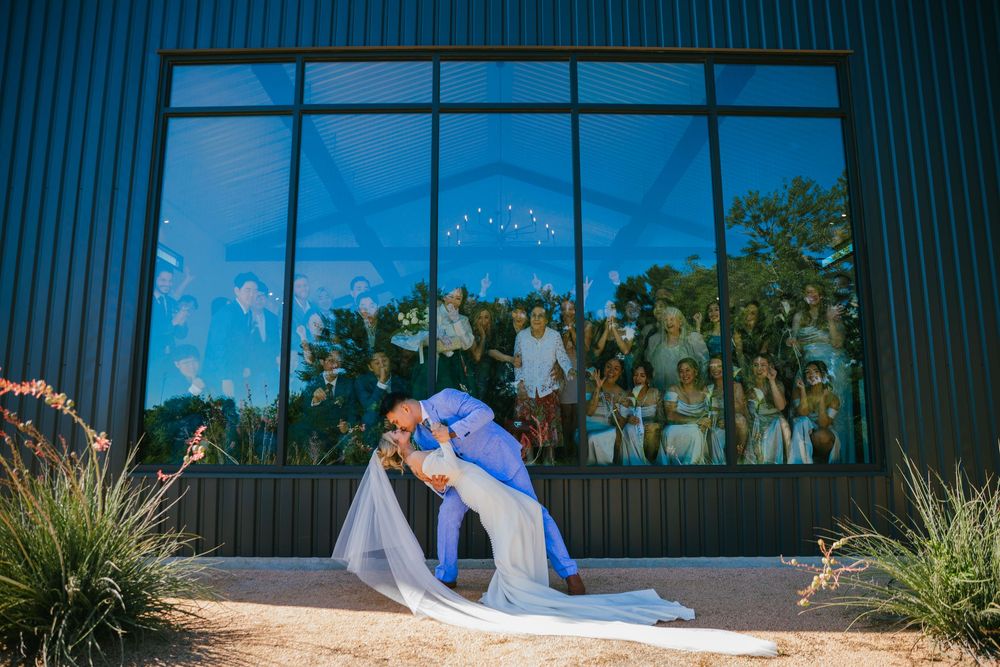 Groom dipping and kissing his bride outdoors in front of a modern glass venue window with cheering guests behind.