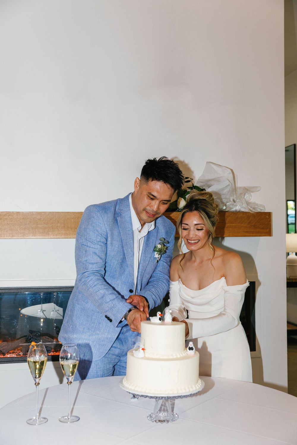 Smiling bride and groom cutting a two-tiered white wedding cake together at an indoor reception table.