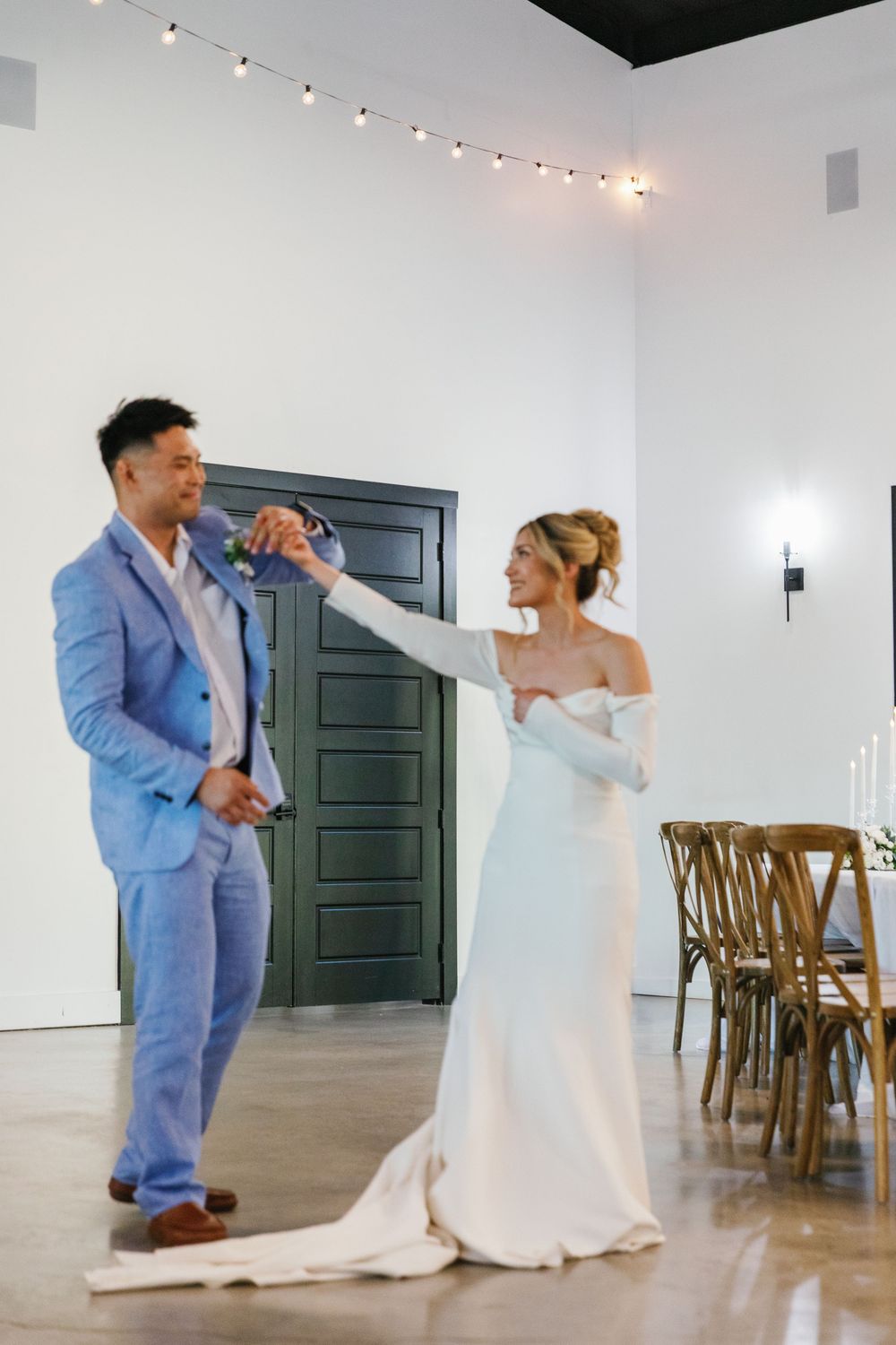 Bride in an off-the-shoulder gown spinning during a first dance with her groom in a light blue suit.