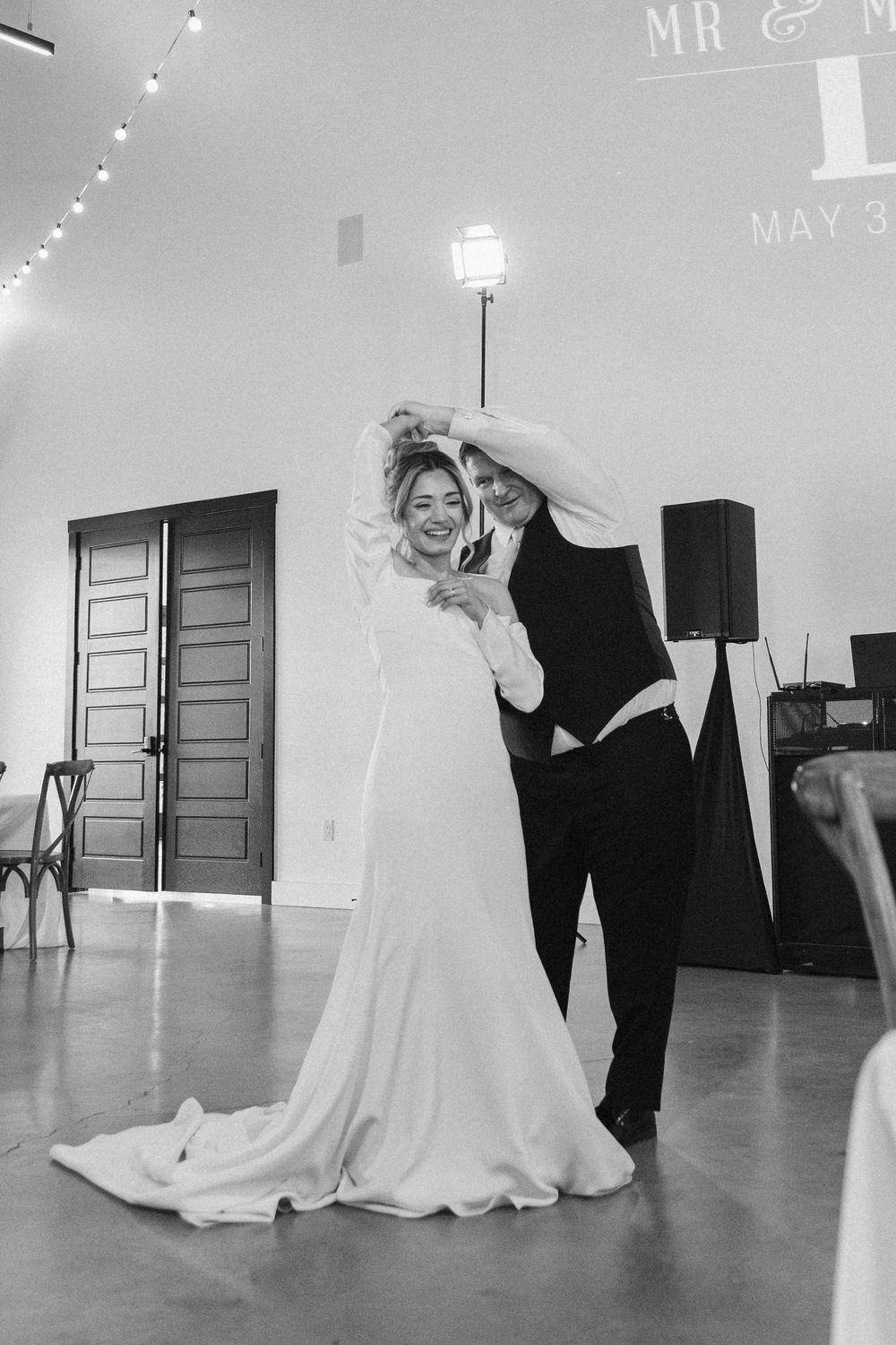 Black and white wedding portrait of a smiling bride being twirled by her father during a reception dance indoors.