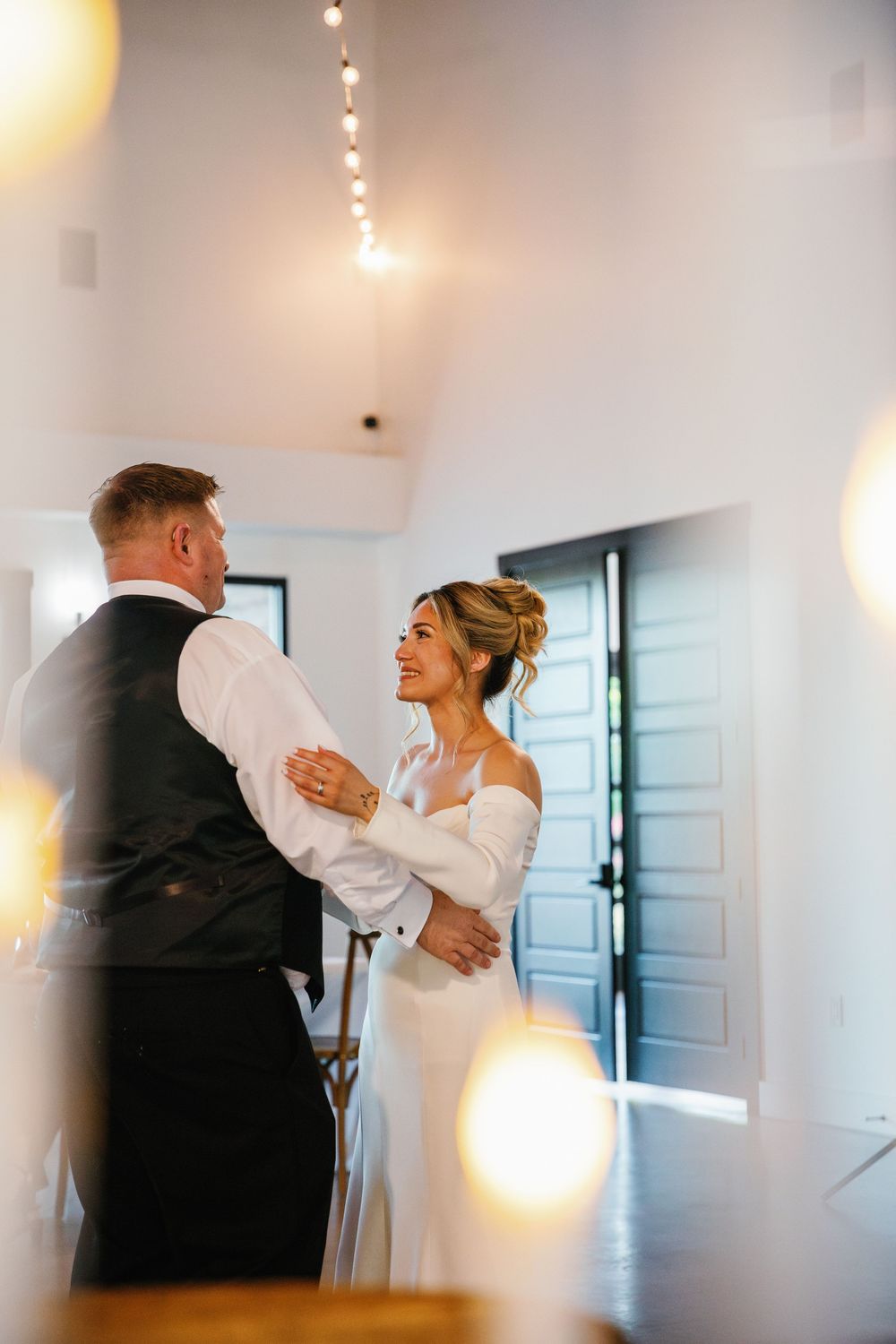 Smiling bride dancing with her father inside a modern indoor wedding venue framed by warm blurred foreground bokeh lights.