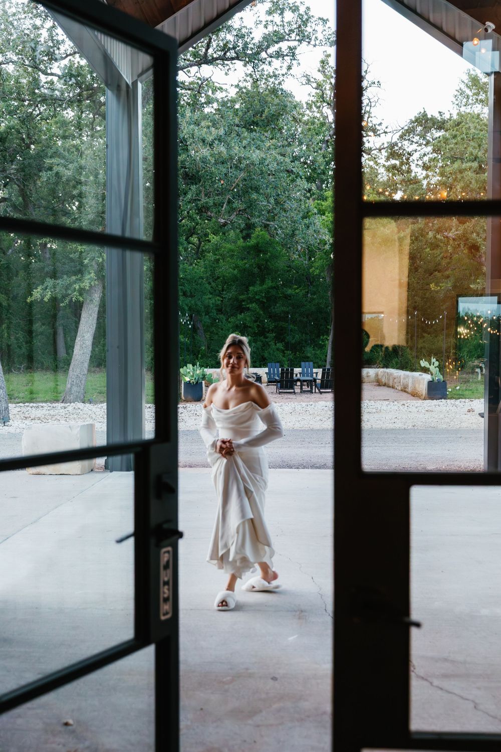 Bride in an off-the-shoulder gown walking toward open glass venue doors during outdoor wedding preparation.