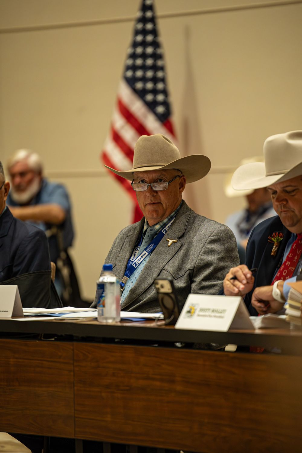 Men in cowboy hats sit at a panel table with an American flag backdrop at a formal meeting.