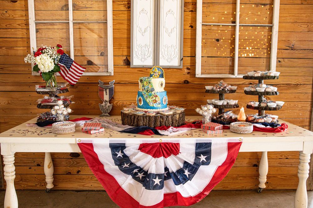 Patriotic dessert table with American flag bunting, tiered cake, and cupcake stands in a rustic barn setting.